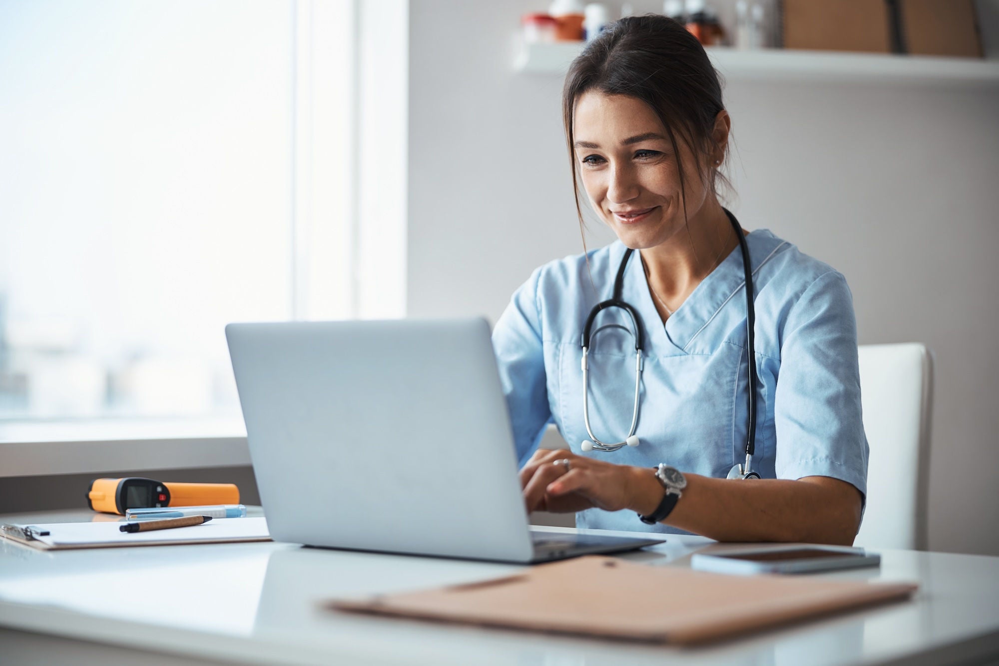 Beautiful young woman physician sitting at the table and smiling while working on modern laptop