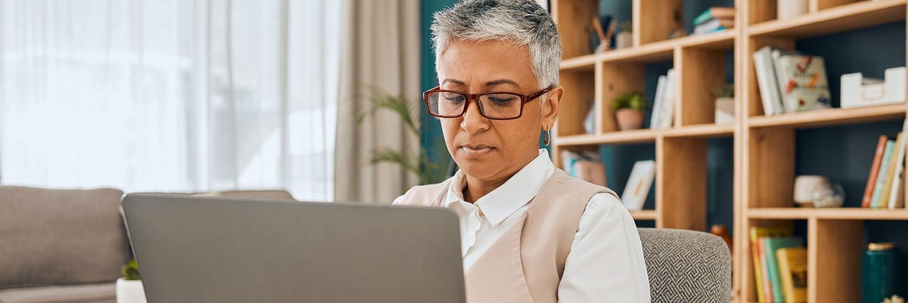 An older adult sits at a desk working intently on a laptop computer. The setting appears to be a cozy home office or living room with bookshelves and decorative items in the background. Soft natural light filters through curtains, creating a calm and focused atmosphere. The person wears glasses and casual-professional clothing while concentrating on the screen.
