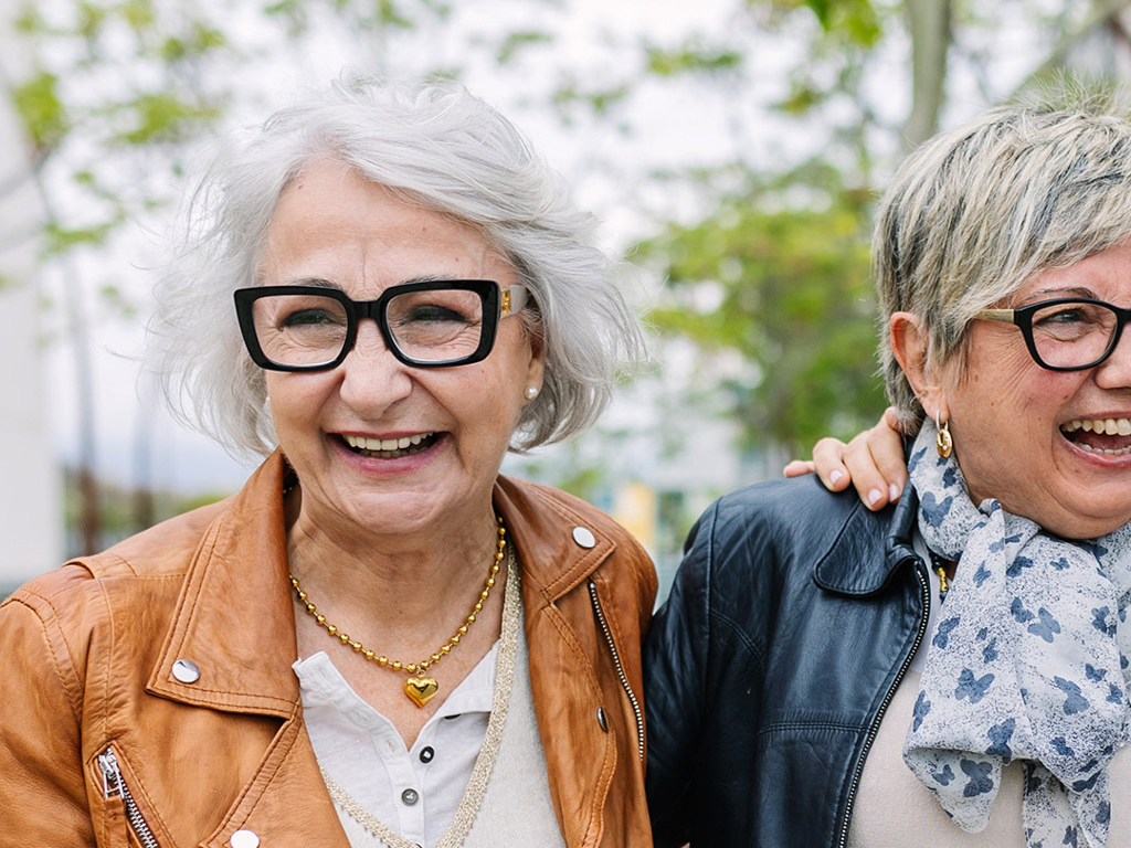 Two older women are pictured standing close together outdoors, each wearing casual jackets. One woman is dressed in a brown leather jacket with a gold necklace, while the other wears a dark jacket and a patterned scarf. The background features greenery and a soft, natural light, suggesting a relaxed and friendly atmosphere. Their body language indicates companionship and warmth.