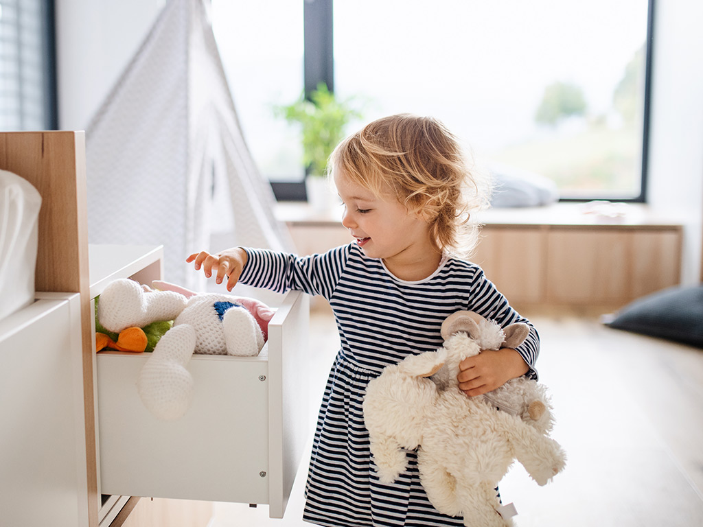 A young child in a striped dress holds plush animals while reaching into a drawer full of soft toys. The scene takes place in a bright, modern bedroom with large windows and minimal furniture. Natural light fills the room, creating a clean and cozy atmosphere. The image conveys playtime, tidiness, and a warm family home environment.