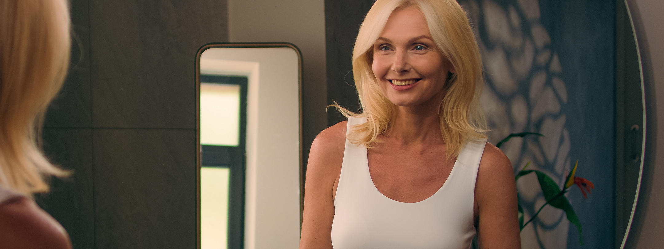 A blonde middle age woman looks in mirror and smiles at herself