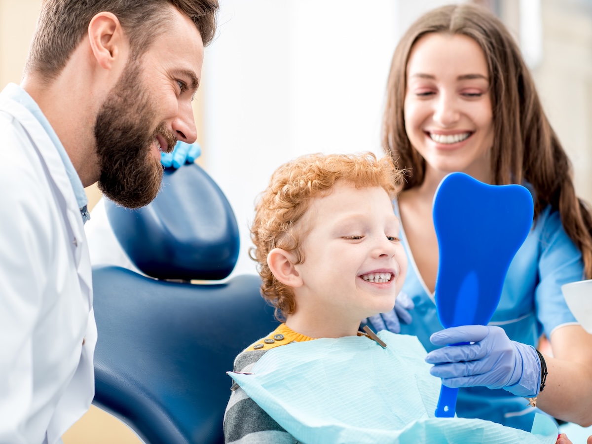 Young boy looking at the mirror with toothy smile sitting on the chair with dentist and assistant at the dental office