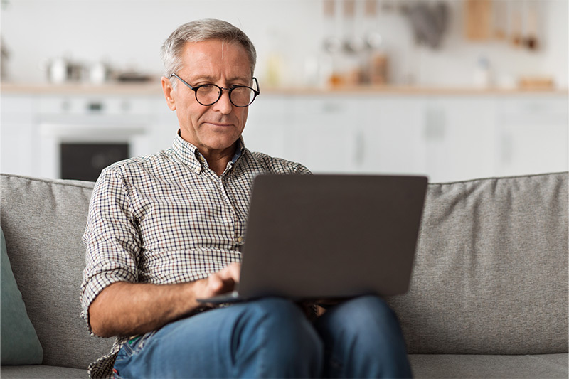 An older man with gray hair and glasses sits on a sofa using a laptop computer. He appears focused on the screen, suggesting remote work, online browsing, or studying. The setting is a cozy home interior with a soft couch and a blurred kitchen in the background. The overall mood is calm, productive, and relaxed.