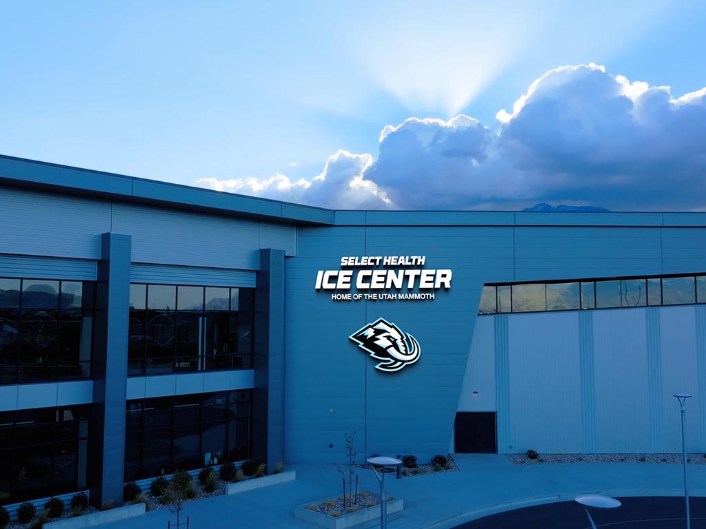 A contemporary ice center building is shown from the outside under a dramatic sky with sun rays breaking through clouds. The facade features large glass windows and smooth blue wall panels. Illuminated signage reads "SELECT HEALTH ICE CENTER" and "HOME OF THE UTAH MAMMOTH" above a stylized mammoth graphic. The surrounding area includes a paved entry space, minimal landscaping, and a calm, empty atmosphere.