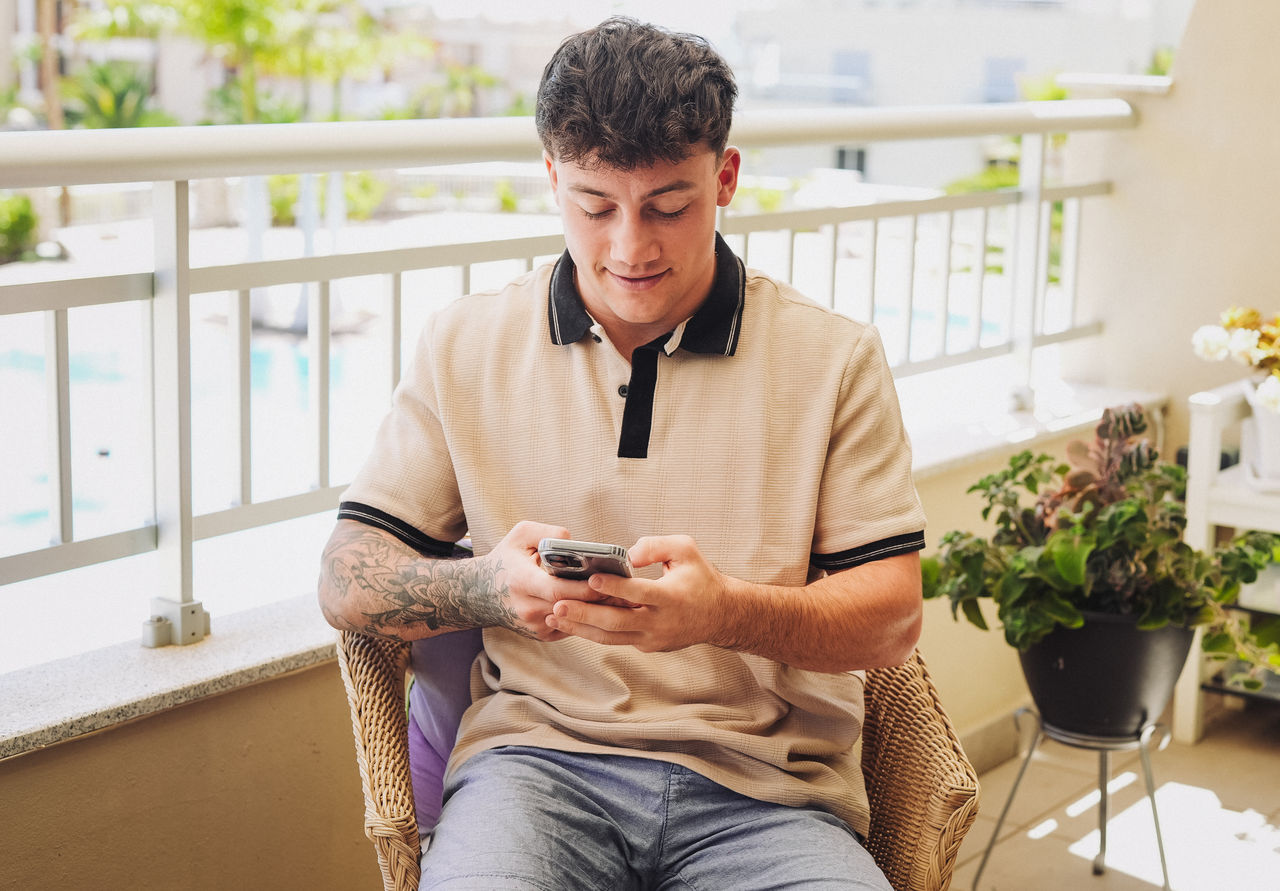 Young caucasian smiling 20 years old boy focused on his smartphone sitting in outdoor terrace enjoying tech and social. Modern lifestyle, relaxation, technology use