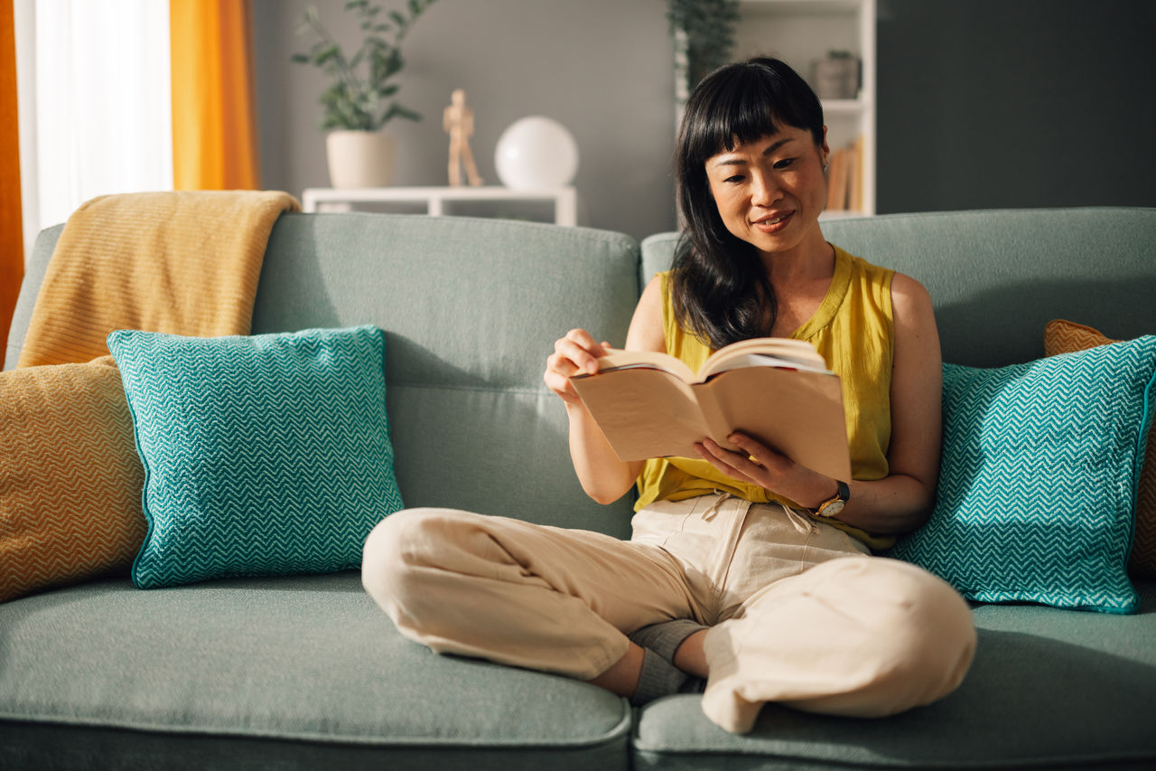 Asian serene woman with a yellow top reads a book while sitting on a cozy green sofa in a well-decorated living room, showcasing her peaceful engagement with literature.