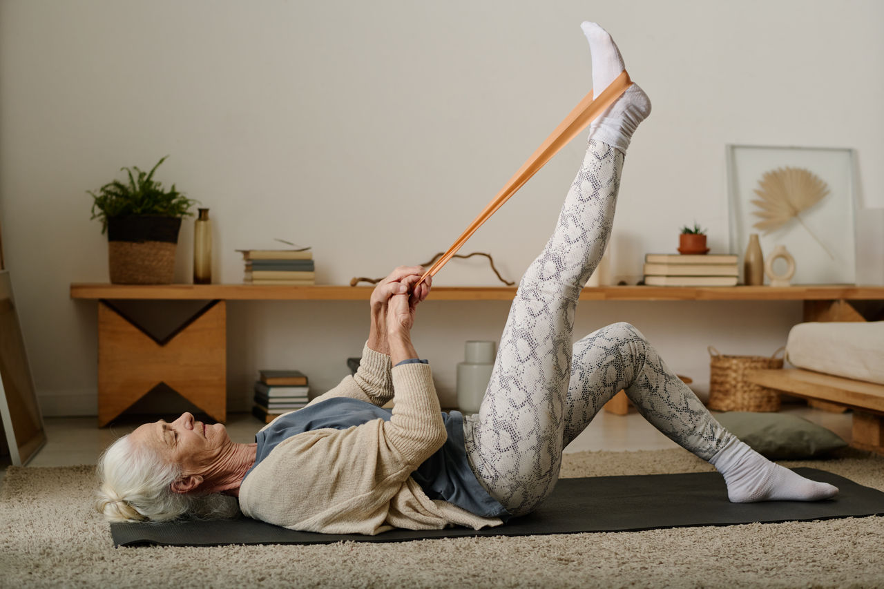 Active senior woman in leggins pulling elastic band while lying on mat in bedroom and raising right leg during physical exercise