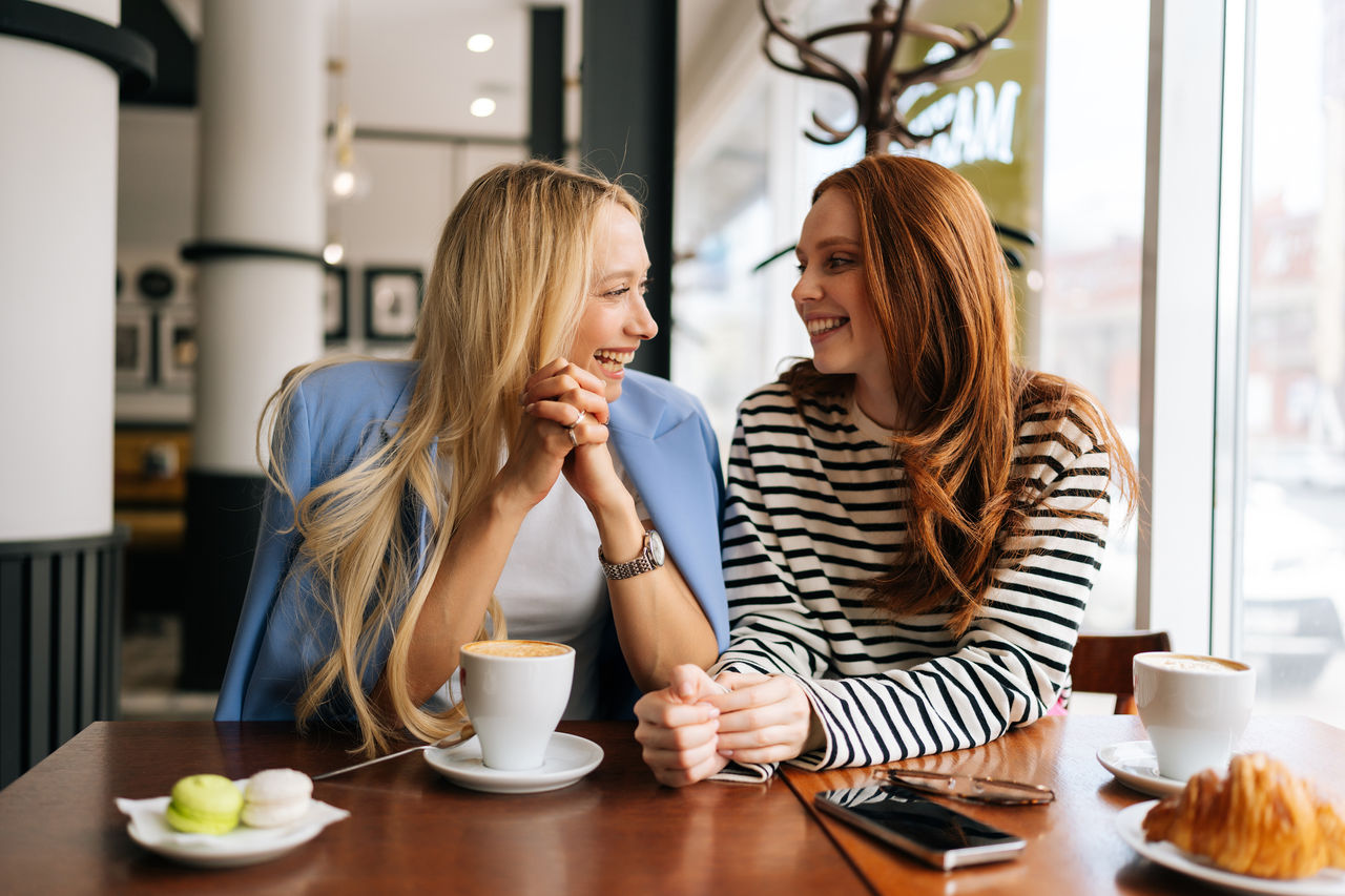 Portrait of laughing blonde female talking to happy redhead woman friend, spending time together during drinking coffee at cozy cafe, sitting at table by window. Concept of woman friendship