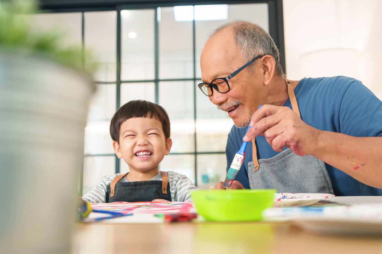 Cheerful senior man and little boy in apron enjoying painting art at home together. Happy Grandfather and Grandchild playing and laughing. 